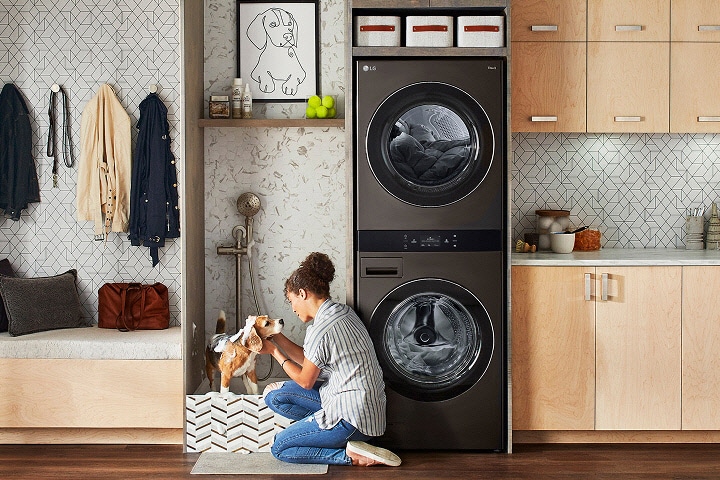 Juodo plieno LG WashTower skalbykloje prieškambaryje su šviesaus medžio spintelėmis, pilkomis plytelėmis išklota sienele, suoliuku ir sieniniais kabliukais.